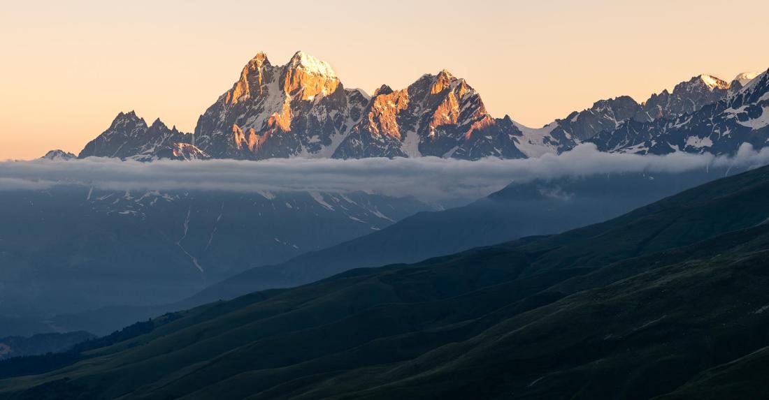 Ušba během východu slunce. Zcela vpravo vystupuje oblý vrchol Elbrusu.